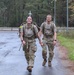 Two military personnel in camouflage uniforms and tactical gear are walking along a wet road through a forested area. Both are carrying large rucksacks and wearing matching unit t-shirts under their gear, suggesting participation in a ruck march or physical training event. The soldier in front is smiling broadly, indicating a positive or accomplished mood.
