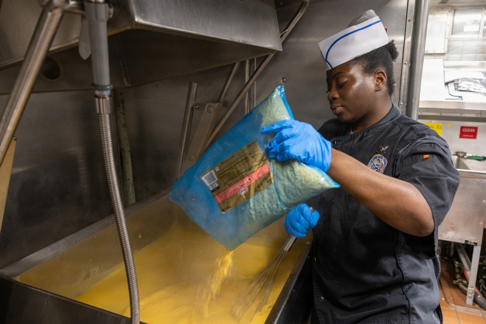 A food service worker wearing a white sailor-style cap and dark uniform is pouring the contents of a large blue bag into a large industrial steam kettle filled with a yellow liquid, likely broth or a sauce. The worker is wearing blue nitrile gloves and appears to be in a large-scale institutional or military kitchen. The setting and uniform suggest this may be a U.S. Navy galley or similar military food service facility.
