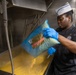 A food service worker wearing a white sailor-style cap and dark uniform is pouring the contents of a large blue bag into a large industrial steam kettle filled with a yellow liquid, likely broth or a sauce. The worker is wearing blue nitrile gloves and appears to be in a large-scale institutional or military kitchen. The setting and uniform suggest this may be a U.S. Navy galley or similar military food service facility.