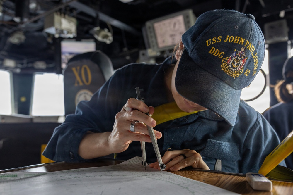 A U.S. Navy sailor aboard the USS John Paul Jones (DDG 53) is using a drafting compass/divider to plot a course on a nautical chart at a navigation table on the ship's bridge. The sailor is wearing a navy blue uniform and a ship-specific ball cap with 'USS John Paul Jones DDG 53' embroidered in gold lettering. In the background, the bridge environment is visible including a chair marked 'XO' (Executive Officer) and various electronic navigation displays.