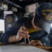A U.S. Navy sailor aboard the USS John Paul Jones (DDG 53) is using a drafting compass/divider to plot a course on a nautical chart at a navigation table on the ship's bridge. The sailor is wearing a navy blue uniform and a ship-specific ball cap with 'USS John Paul Jones DDG 53' embroidered in gold lettering. In the background, the bridge environment is visible including a chair marked 'XO' (Executive Officer) and various electronic navigation displays.