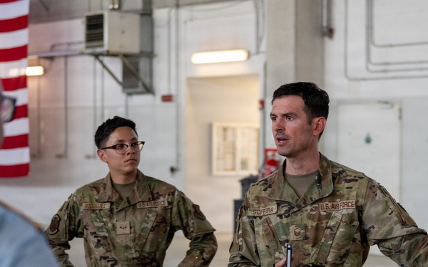 Two U.S. Air Force personnel in OCP (Operational Camouflage Pattern) uniforms are engaged in a briefing or discussion inside what appears to be a military hangar or large facility. The individual on the right, whose name tape reads 'SOFFERA', is actively speaking and gesturing with outstretched arms while holding a small device. A third person in a light blue uniform is visible from behind in the foreground.