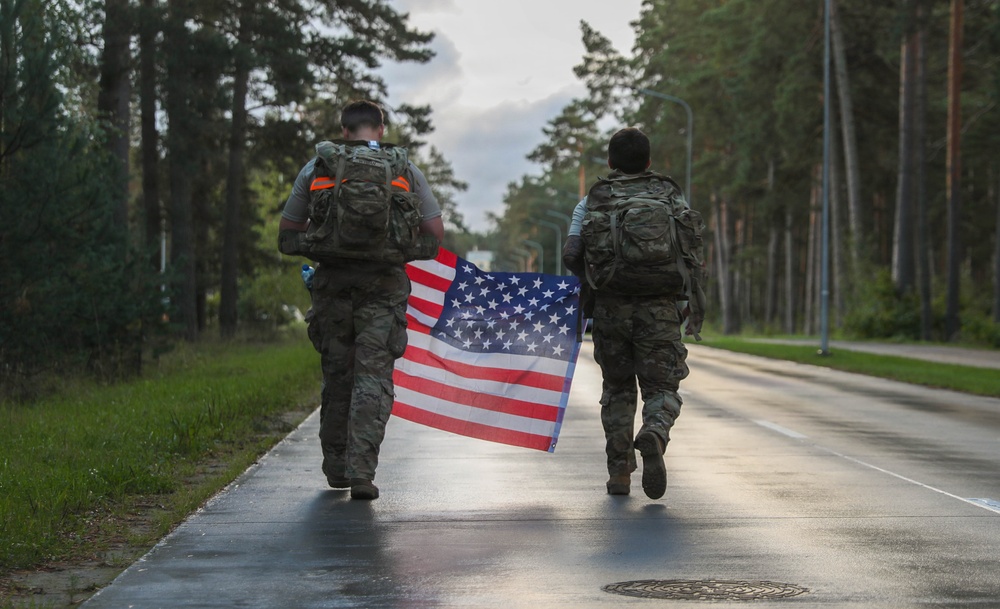 Two military personnel in camouflage uniforms and tactical backpacks are walking away from the camera along a wet paved road lined with tall trees. They are jointly carrying an American flag between them. The setting appears to be a military base or installation, likely during or after a ruck march or commemorative event.