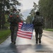 Two military personnel in camouflage uniforms and tactical backpacks are walking away from the camera along a wet paved road lined with tall trees. They are jointly carrying an American flag between them. The setting appears to be a military base or installation, likely during or after a ruck march or commemorative event.