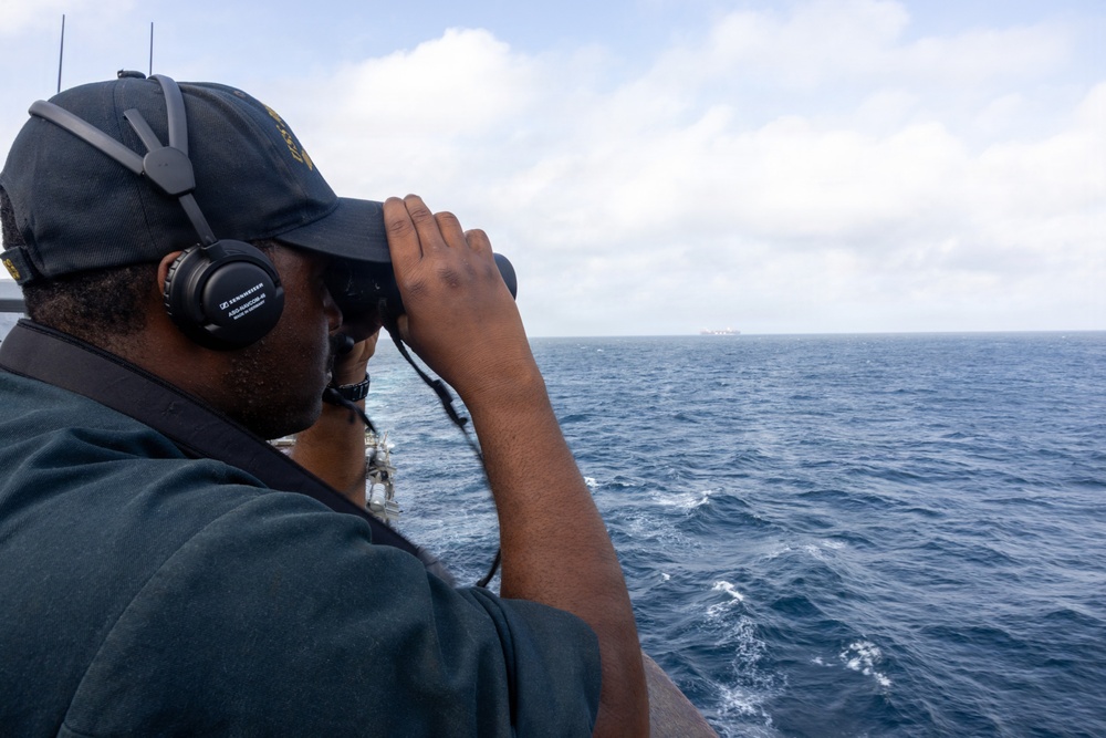 A person wearing a dark baseball cap and Sennheiser headphones is using binoculars to observe the open ocean from what appears to be a naval or military vessel. In the distant horizon, a large ship or vessel is faintly visible. The scene suggests maritime surveillance or watch-keeping duties.