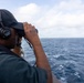 A person wearing a dark baseball cap and Sennheiser headphones is using binoculars to observe the open ocean from what appears to be a naval or military vessel. In the distant horizon, a large ship or vessel is faintly visible. The scene suggests maritime surveillance or watch-keeping duties.