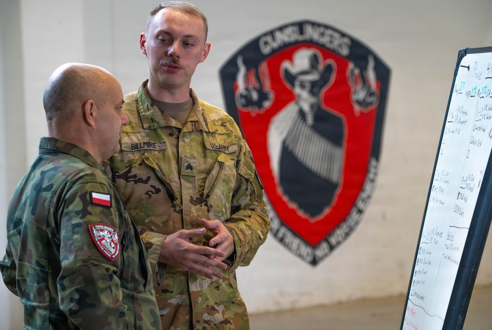 Two military personnel are engaged in a briefing or discussion near a whiteboard with written notes. One soldier wears a US Army uniform with the name tape 'BILLMIRE' and Sergeant rank insignia, while the other wears a Polish military camouflage uniform with a Polish flag patch. They are standing in front of a unit insignia mural reading 'GUNSLINGERS' with the motto 'NO FRIEND NO WORSE ENEMY'.
