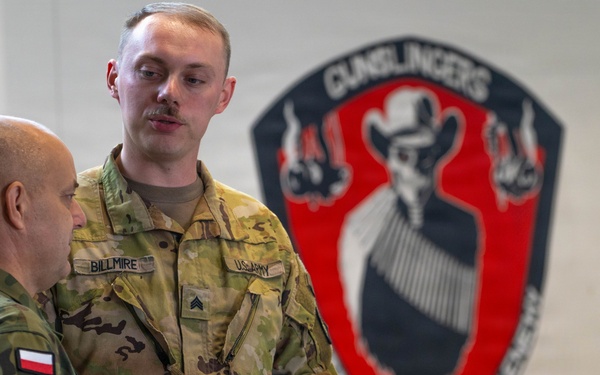 Two military personnel are engaged in a briefing or discussion near a whiteboard with written notes. One soldier wears a US Army uniform with the name tape 'BILLMIRE' and Sergeant rank insignia, while the other wears a Polish military camouflage uniform with a Polish flag patch. They are standing in front of a unit insignia mural reading 'GUNSLINGERS' with the motto 'NO FRIEND NO WORSE ENEMY'.