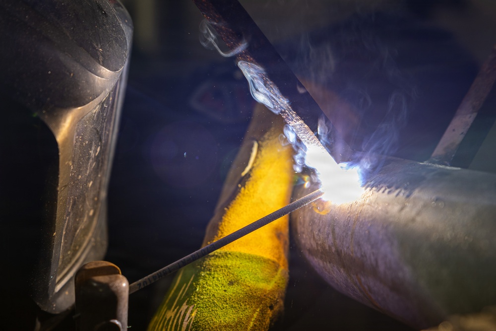 A welder is actively welding a metal pipe or cylindrical workpiece, with a bright arc visible at the point of contact. The worker is wearing a welding helmet and yellow protective gloves, with visible smoke and sparks emanating from the weld zone. The close-up shot captures the intense light and heat of the welding process in a dark industrial environment.