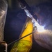 A welder is actively welding a metal pipe or cylindrical workpiece, with a bright arc visible at the point of contact. The worker is wearing a welding helmet and yellow protective gloves, with visible smoke and sparks emanating from the weld zone. The close-up shot captures the intense light and heat of the welding process in a dark industrial environment.