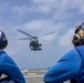 Two naval personnel in blue jerseys and white helmets with hearing protection observe a military helicopter approaching or departing from a ship's flight deck. The helicopter, consistent with an MH-60 Seahawk variant, is captured mid-flight with rotor blades in motion. The scene depicts standard naval aviation flight deck operations.