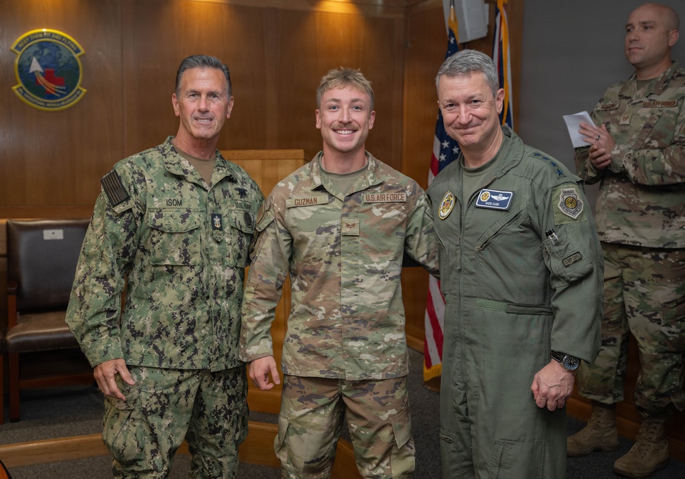 Three military personnel pose for a formal photo in what appears to be an official military building or conference room. The individuals represent different branches of the U.S. military, including the U.S. Navy and U.S. Air Force, with name tapes visible on their uniforms. A fourth service member stands in the background holding a document.
