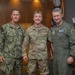 Three military personnel pose for a formal photo in what appears to be an official military building or conference room. The individuals represent different branches of the U.S. military, including the U.S. Navy and U.S. Air Force, with name tapes visible on their uniforms. A fourth service member stands in the background holding a document.