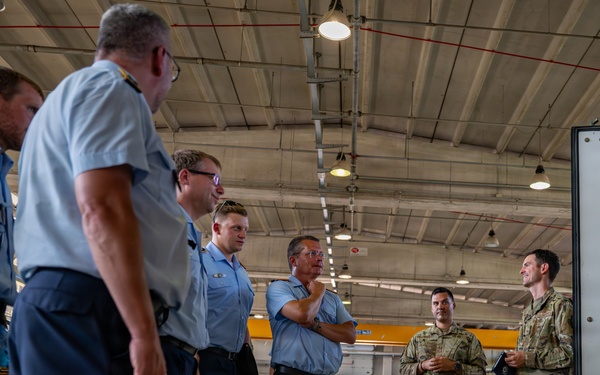 A group of military personnel in two distinct uniforms are gathered inside what appears to be a military hangar or maintenance facility. Several individuals wearing light blue uniforms are facing two personnel in camouflage (OCP/MultiCam) uniforms, suggesting a briefing or tour is taking place. The setting indicates an inter-service or multinational military exchange or visit.