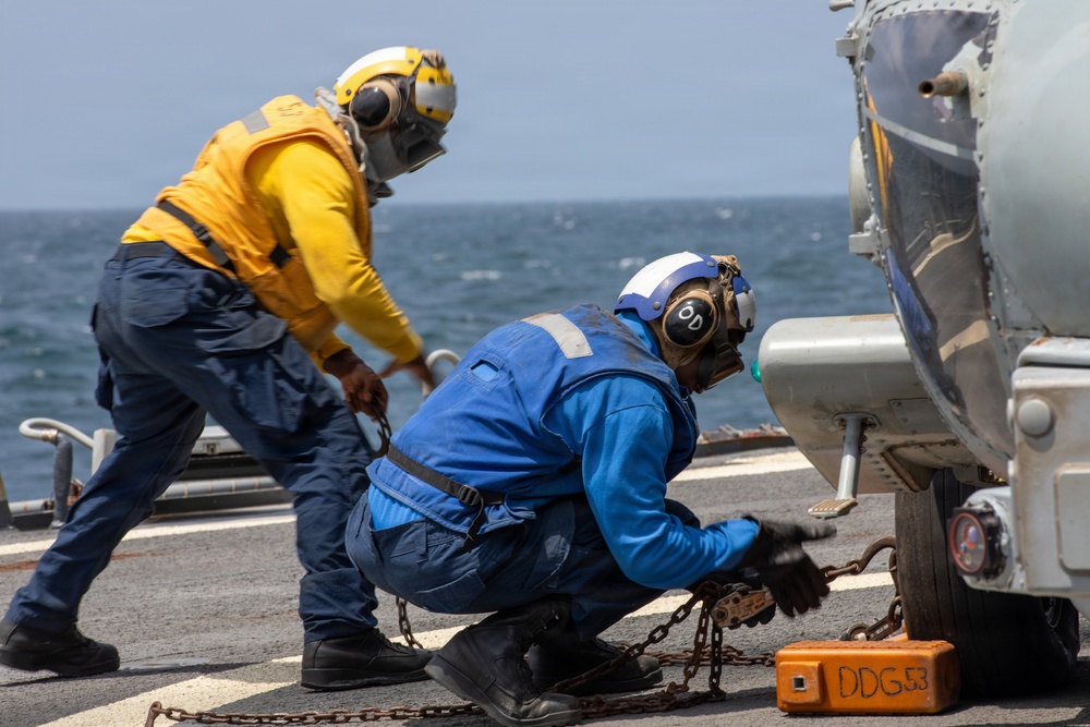 Two naval personnel are working on a ship's flight deck, securing what appears to be a helicopter with chains and tie-downs. One sailor is wearing a yellow jersey and yellow helmet, while the other wears a blue jersey and white helmet, indicating different crew roles. A visible marking 'DDG 53' on an orange chock identifies the vessel as USS John Paul Jones, a U.S. Navy Arleigh Burke-class destroyer.