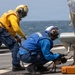 Two naval personnel are working on a ship's flight deck, securing what appears to be a helicopter with chains and tie-downs. One sailor is wearing a yellow jersey and yellow helmet, while the other wears a blue jersey and white helmet, indicating different crew roles. A visible marking 'DDG 53' on an orange chock identifies the vessel as USS John Paul Jones, a U.S. Navy Arleigh Burke-class destroyer.