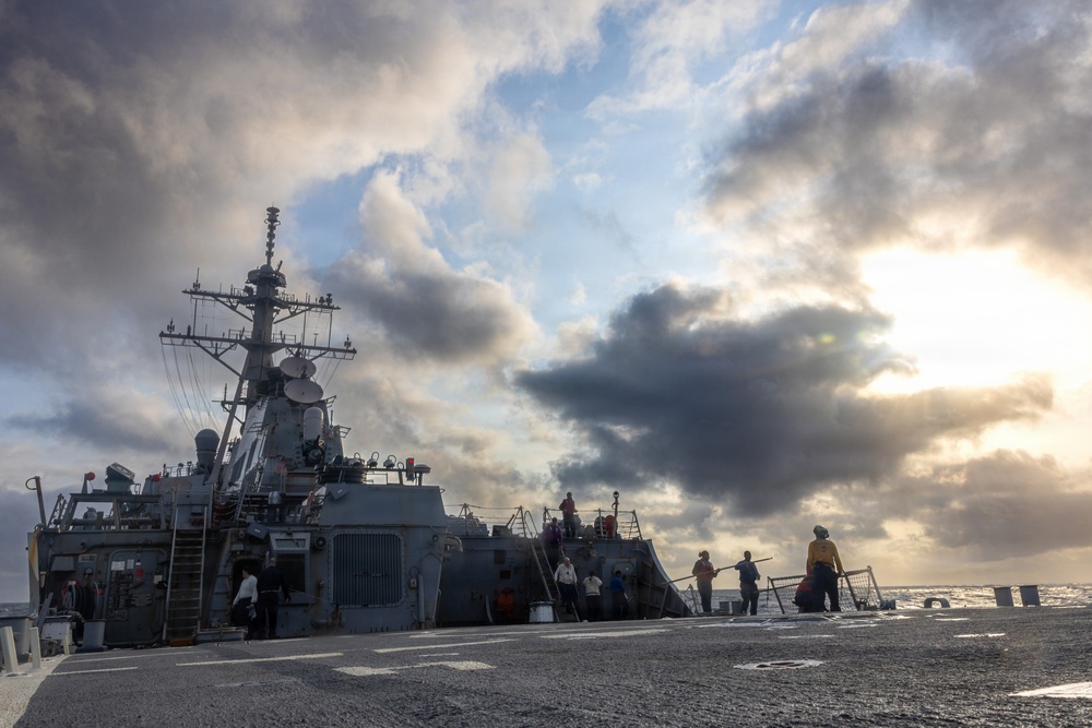 A U.S. Navy warship, consistent with an Arleigh Burke-class guided-missile destroyer, is photographed from its deck level with crew members visible on the exterior. The vessel is at sea under a dramatic, partially overcast sky with sunlight breaking through the clouds. Multiple sailors appear to be conducting deck operations or maintenance activities.