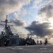 A U.S. Navy warship, consistent with an Arleigh Burke-class guided-missile destroyer, is photographed from its deck level with crew members visible on the exterior. The vessel is at sea under a dramatic, partially overcast sky with sunlight breaking through the clouds. Multiple sailors appear to be conducting deck operations or maintenance activities.
