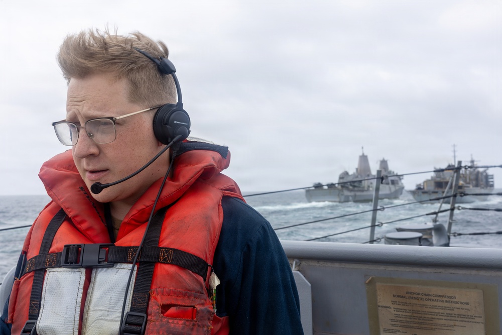 A naval personnel member wearing a red life vest and communications headset stands on the deck of a military vessel at sea. In the background, at least two large naval warships are visible navigating rough waters under an overcast sky. The scene appears to depict an underway replenishment or fleet formation operation.