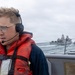 A naval personnel member wearing a red life vest and communications headset stands on the deck of a military vessel at sea. In the background, at least two large naval warships are visible navigating rough waters under an overcast sky. The scene appears to depict an underway replenishment or fleet formation operation.