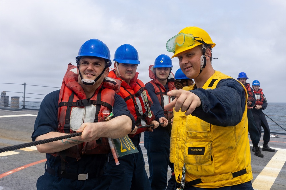A group of naval personnel are conducting a line-handling or replenishment-at-sea (RAS) drill on the deck of a military vessel. Several sailors wearing blue hard hats and orange life vests are pulling a rope, while a supervisor in a yellow hard hat, yellow safety vest, and communication headset points and provides direction. The overcast sky and open ocean are visible in the background.