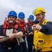 A group of naval personnel are conducting a line-handling or replenishment-at-sea (RAS) drill on the deck of a military vessel. Several sailors wearing blue hard hats and orange life vests are pulling a rope, while a supervisor in a yellow hard hat, yellow safety vest, and communication headset points and provides direction. The overcast sky and open ocean are visible in the background.
