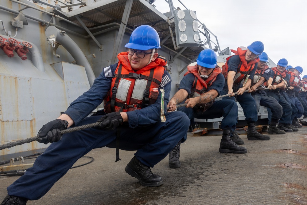 A line of naval personnel are pulling a heavy rope in unison on the deck of a military vessel. The sailors are wearing standard navy working uniforms, blue hard hats, and orange life vests, suggesting a mooring or line-handling evolution. The image is taken from a low angle, emphasizing the physical effort and coordinated teamwork of the crew.