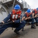 A line of naval personnel are pulling a heavy rope in unison on the deck of a military vessel. The sailors are wearing standard navy working uniforms, blue hard hats, and orange life vests, suggesting a mooring or line-handling evolution. The image is taken from a low angle, emphasizing the physical effort and coordinated teamwork of the crew.