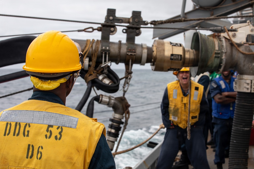 Naval personnel are conducting a replenishment-at-sea (RAS) operation aboard a U.S. Navy destroyer. The foreground sailor's vest is stenciled with 'DDG 53 D6', identifying the vessel as USS John Paul Jones (DDG-53). Crew members in yellow safety vests and hard hats are managing fuel or supply transfer hoses and rigging between ships at sea.