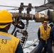 Naval personnel are conducting a replenishment-at-sea (RAS) operation aboard a U.S. Navy destroyer. The foreground sailor's vest is stenciled with 'DDG 53 D6', identifying the vessel as USS John Paul Jones (DDG-53). Crew members in yellow safety vests and hard hats are managing fuel or supply transfer hoses and rigging between ships at sea.