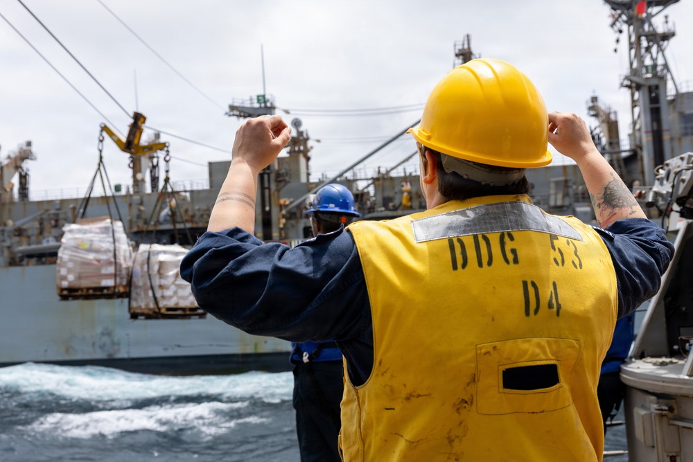 A U.S. Navy sailor wearing a yellow safety vest marked 'DDG 53 D4' and a yellow hard hat signals during an underway replenishment (UNREP) operation at sea. Palletized cargo is being transferred via highline between two vessels. A second crew member in a blue hard hat is visible in the background observing the operation.