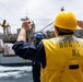A U.S. Navy sailor wearing a yellow safety vest marked 'DDG 53 D4' and a yellow hard hat signals during an underway replenishment (UNREP) operation at sea. Palletized cargo is being transferred via highline between two vessels. A second crew member in a blue hard hat is visible in the background observing the operation.