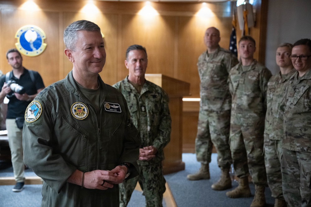 A military gathering in what appears to be an official briefing or ceremony room, featuring personnel from multiple U.S. military branches. The foreground figure wears an olive green flight suit with patches including what appears to be a Joint Chiefs of Staff insignia. Personnel in Navy blues and Army/Air Force multicam uniforms are visible in the background, along with a civilian photographer.