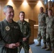 A military gathering in what appears to be an official briefing or ceremony room, featuring personnel from multiple U.S. military branches. The foreground figure wears an olive green flight suit with patches including what appears to be a Joint Chiefs of Staff insignia. Personnel in Navy blues and Army/Air Force multicam uniforms are visible in the background, along with a civilian photographer.