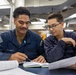 Two U.S. Navy sailors in navy blue utility uniforms are seated at a table reviewing documents and a notebook together. The setting appears to be an interior workspace aboard a naval vessel, indicated by the industrial overhead piping, ducting labeled 'RECIRC' and 'SUPPLY', and fluorescent lighting. The sailors appear to be engaged in a study or training session.