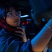 A person wearing clear-framed glasses and a dark uniform holds a red telephone handset in what appears to be a dimly lit military operations or command center. The environment is illuminated by blue ambient lighting with red indicator lights visible in the background. A second individual is partially visible in the background, also in a dark uniform.