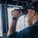 A U.S. Navy sailor is standing watch on the bridge of a naval vessel, using binoculars to scan the horizon. The sailor is wearing standard Navy working uniform (NWU) in navy blue, along with a command ball cap bearing a U.S. flag patch and partial hull designation 'DDG', indicating a destroyer-class ship. The scene appears to be taken from inside the bridge wing or pilothouse with an open view to the sea.