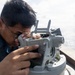 A U.S. Navy sailor is using a gyrocompass or bearing circle (alidade) mounted on the bridge wing of a naval vessel at sea. A second sailor wearing a headset and cover (cap) is visible in the background. The setting appears to be an open-ocean environment under bright, hazy conditions.
