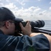 A man wearing a dark baseball cap and glasses is photographing over open water using a camera equipped with a large telephoto lens. He is positioned low, resting the camera on what appears to be a railing or ledge of a boat. The background shows a calm ocean horizon under a partly cloudy sky.