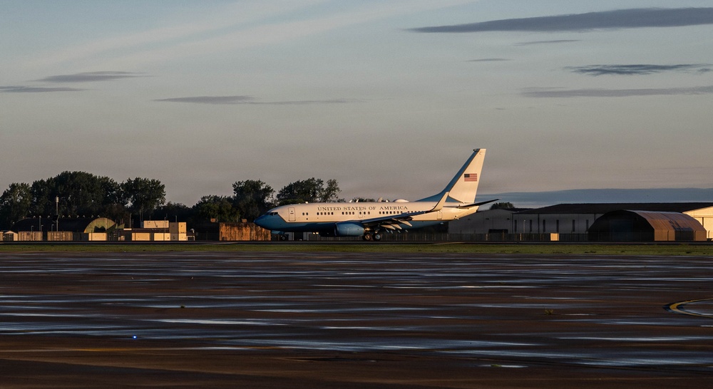 A United States of America government aircraft, consistent with an Air Force executive transport, is parked on a tarmac at what appears to be a military airfield. The aircraft features the distinctive blue and white livery with 'UNITED STATES OF AMERICA' lettering and an American flag on the tail. The image is taken during golden hour (early morning or late evening), with hangars and trees visible in the background.