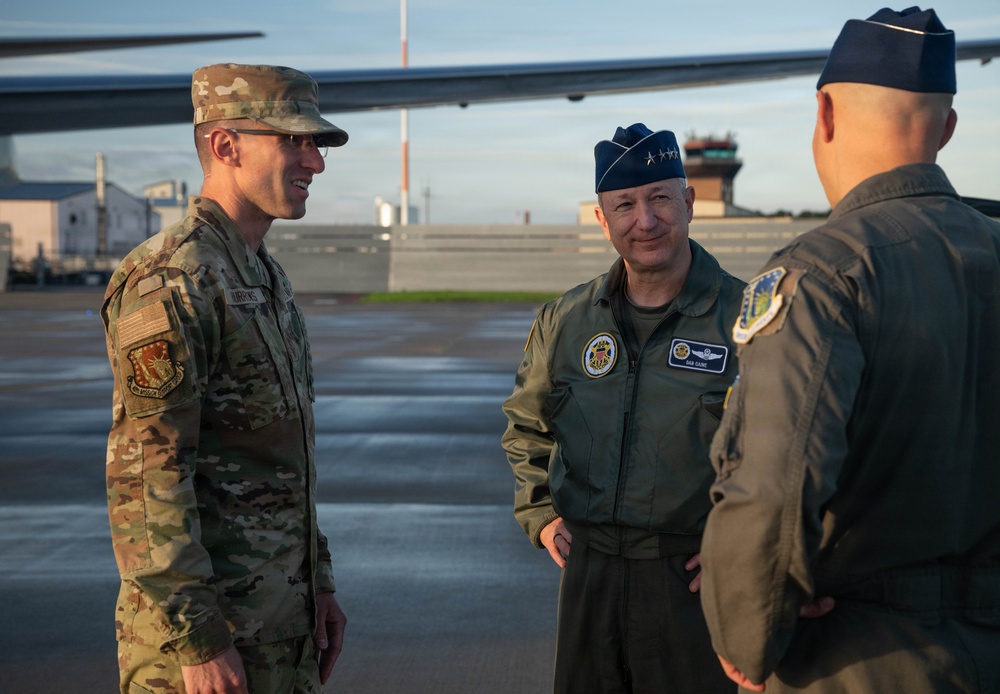 Three U.S. Air Force personnel are conversing on an aircraft flight line, likely at a military air base. The central figure wears a service dress flight cap with four-star rank insignia and a name tag reading 'DAN CAINE,' indicating a very senior officer. The setting appears to be during early morning or evening hours based on the golden lighting conditions.