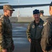 Three U.S. Air Force personnel are conversing on an aircraft flight line, likely at a military air base. The central figure wears a service dress flight cap with four-star rank insignia and a name tag reading 'DAN CAINE,' indicating a very senior officer. The setting appears to be during early morning or evening hours based on the golden lighting conditions.