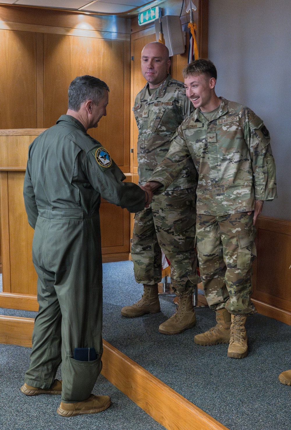 A U.S. Air Force officer in an olive green flight suit is shaking hands with a uniformed airman identified by a name tape reading 'GUZMAN,' while a second airman in OCP (Operational Camouflage Pattern) uniform stands nearby. The setting appears to be an official indoor military facility with wood-paneled walls and a flag in the background. The interaction suggests a formal recognition or greeting ceremony.