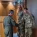 A U.S. Air Force officer in an olive green flight suit is shaking hands with a uniformed airman identified by a name tape reading 'GUZMAN,' while a second airman in OCP (Operational Camouflage Pattern) uniform stands nearby. The setting appears to be an official indoor military facility with wood-paneled walls and a flag in the background. The interaction suggests a formal recognition or greeting ceremony.