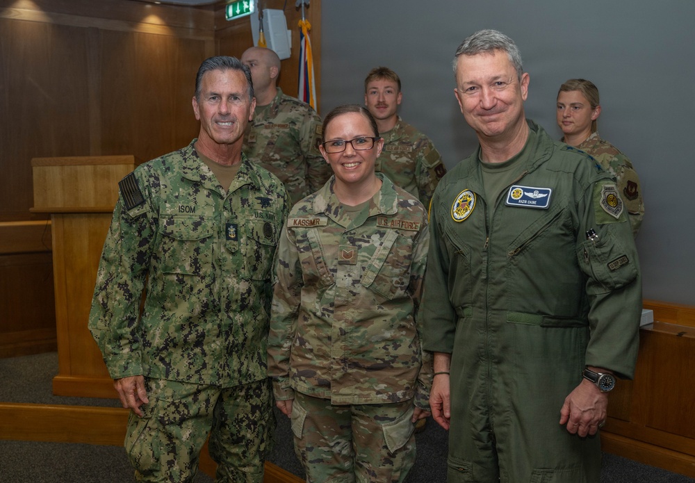 Three military personnel pose for a formal photo in what appears to be an indoor military facility. The individuals represent different branches of the U.S. Armed Forces, including the U.S. Navy and U.S. Air Force, identifiable by their distinct uniforms and name tapes. Additional uniformed personnel are visible in the background.