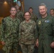 Three military personnel pose for a formal photo in what appears to be an indoor military facility. The individuals represent different branches of the U.S. Armed Forces, including the U.S. Navy and U.S. Air Force, identifiable by their distinct uniforms and name tapes. Additional uniformed personnel are visible in the background.