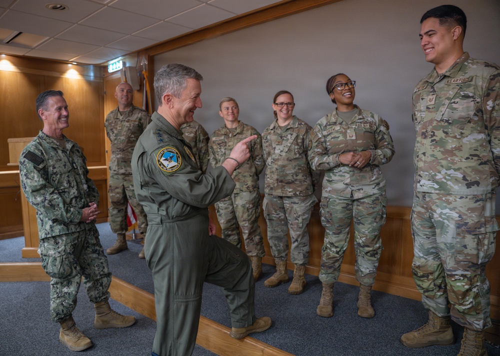 A group of U.S. military personnel from multiple branches are gathered in what appears to be an indoor briefing or meeting room. A senior officer in an olive green flight suit is gesturing and speaking animatedly to a line of Air Force personnel in OCP (Operational Camouflage Pattern) uniforms, while a Navy officer in NWU (Navy Working Uniform) stands to the left smiling. The atmosphere appears relaxed and positive, suggesting an informal interaction or recognition event.