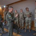 A group of U.S. military personnel from multiple branches are gathered in what appears to be an indoor briefing or meeting room. A senior officer in an olive green flight suit is gesturing and speaking animatedly to a line of Air Force personnel in OCP (Operational Camouflage Pattern) uniforms, while a Navy officer in NWU (Navy Working Uniform) stands to the left smiling. The atmosphere appears relaxed and positive, suggesting an informal interaction or recognition event.