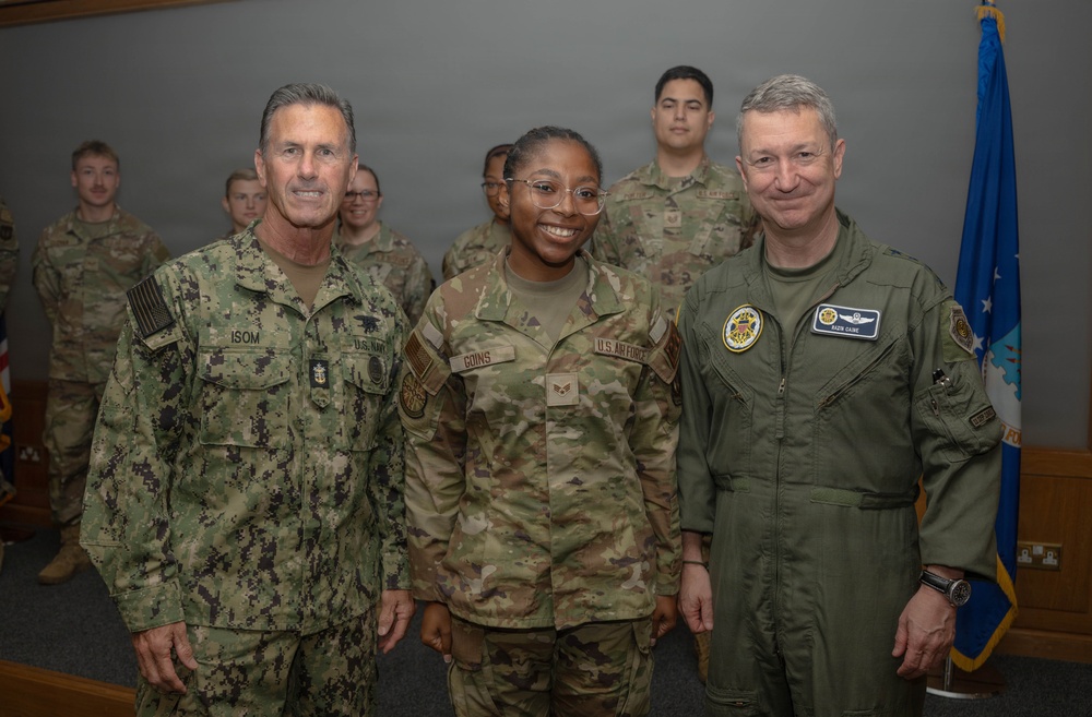 Three military personnel pose for a formal photograph in what appears to be an indoor military facility. The central figure, identified by name tape as 'GOINS' of the U.S. Air Force, is flanked by a U.S. Navy officer named 'ISOM' on the left and an Air Force officer in a flight suit on the right. Several additional uniformed personnel are visible in the background.