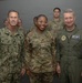 Three military personnel pose for a formal photograph in what appears to be an indoor military facility. The central figure, identified by name tape as 'GOINS' of the U.S. Air Force, is flanked by a U.S. Navy officer named 'ISOM' on the left and an Air Force officer in a flight suit on the right. Several additional uniformed personnel are visible in the background.